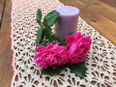 Cremefarbener gehäkelter Tischläufer mit Blütenmuster, pinke Rosen und pastelllila Kerze auf Holztisch