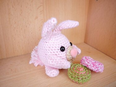 Rosa gehäkeltes Amigurumi-Häschen mit schwarzen Augen und zwei gehäkelten Dekorationen auf Holzuntergrund