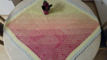 Quadratisches handgehäkeltes Deckchen mit Farbverlauf von Gelb zu Pink und hellem Wellenrand auf einem Tisch, dekoriert mit einem kleinen Blumentopf.