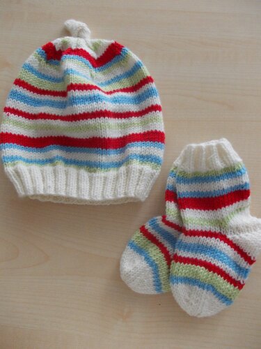 Gestricktes Babyset mit Ringelstreifen: Mütze mit kleinem Knoten und Söckchen in Rot, Hellblau, Hellgrün und Creme auf hellem Holzuntergrund.