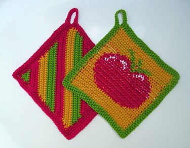 Zwei gehäkelte Topflappen in Pink, Gelb und Grün; einer mit Apfelmotiv, der andere gestreift.