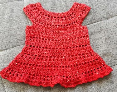 Rotes gehäkeltes Babykleid mit offenem Muster und dezenten Glitzereffekten auf hellem Hintergrund.