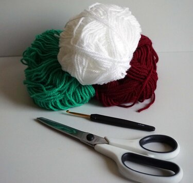 Green, white and red yarn with a crochet hook and scissors on a white surface.