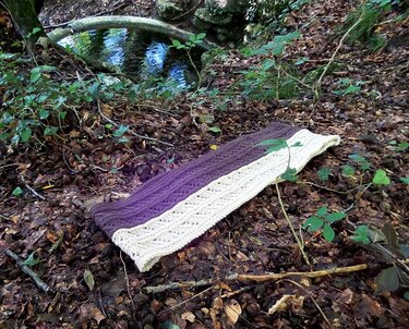 Purple and cream cable and lace knitted loop scarf laid flat on leaf-strewn forest floor