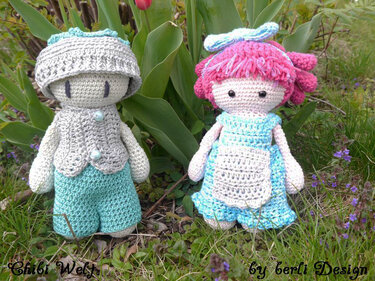 Zwei gehäkelte Amigurumi-Puppen, eine mit grauer Mütze und türkisfarbener Hose, eine mit rosa Haaren und hellblauem Kleid, platziert im Gras vor Tulpen.