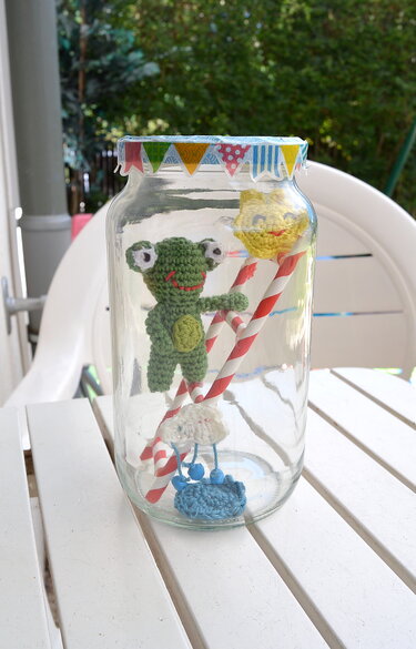 Gehäkelter grüner Frosch, gelbe Sonne und Wolke mit Tropfen in einem Glas mit rot-weiß gestreiften Strohhalmen.