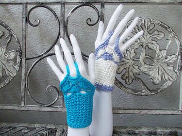 Zwei gehäkelte fingerlose Handschuhe in Türkis und Weiß-Lila auf Mannequin-Händen vor ornamentalem Metallgitter