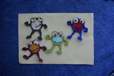 Vier gehäkelte Frosch-Applikationen in unterschiedlichen Farben auf hellem Papier vor blauem Handtuch
