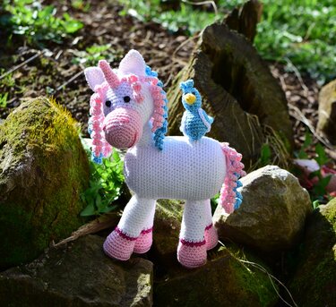 Gehäkeltes weißes Einhorn mit rosa-blauer Mähne und pinken Hufen, mit kleinem blauen Häkelvogel auf dem Rücken, auf moosbewachsenen Felsen