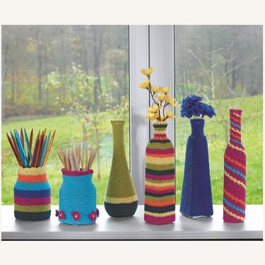 Gehäkelte Hüllen und Behälter in bunten Streifen- und Uni-Farben auf einer Fensterbank, gefüllt mit Stiften und Stricknadeln.