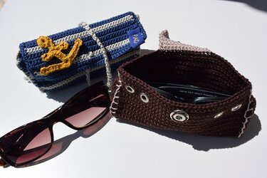 Zwei gehäkelte Brillenetuis: blau-weiß gestreift mit Anker und braun mit Metallösen; eine Sonnenbrille liegt daneben.