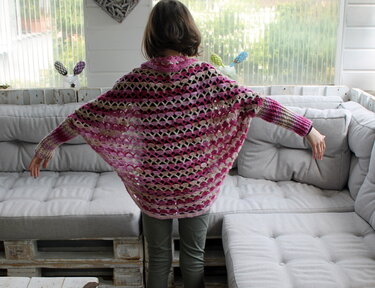 Rückenansicht einer Person trägt gehäkelten Seelenwärmer in Rosa- und Naturtönen mit offenem Muschelmuster und langen Bündchen vor hellem Sofa