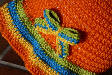 Nahaufnahme einer gehäkelten Kindermütze in Orange mit gelben, blauen und grünen Streifen sowie einer kleinen Schleife.