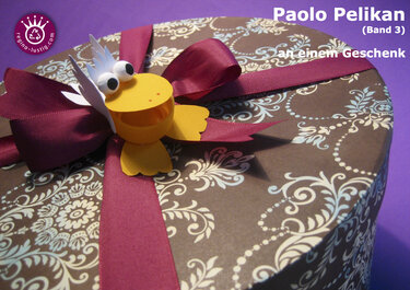 Runde gemusterte Geschenkbox mit weinroter Schleife und gelbem Papierpelikan mit Wackelaugen