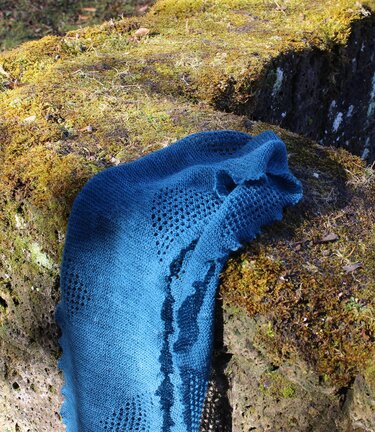 Petrolblauer gestrickter Schal mit Lochmuster und wellenförmiger Kante auf moosigem Stein.