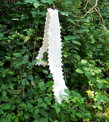 Long white knitted scarf with scalloped edge hanging on green foliage