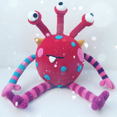 Rotes gehäkeltes Amigurumi-Monster mit drei Augenstielen, türkisfarbenen Punkten und gestreiften Armen und Beinen auf hellem Hintergrund
