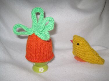 Orange gestrickter Karotten-Eierwärmer mit grünen Blättern auf Eierbecher, daneben gelbes gestricktes Küken