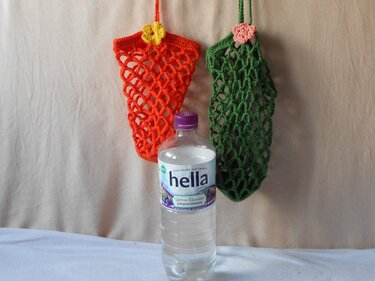 Zwei gehäkelte Netzbeutel, einer orange mit gelber Blüte und einer grün mit rosafarbener Blüte; eine durchsichtige Flasche steht davor