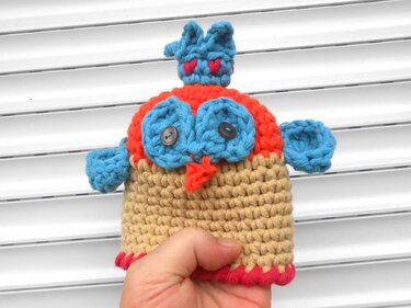 Hand hält gehäkelte Mini-Eule aus beigem, blauem und orangefarbenem Garn mit Knopfaugen vor weißer Jalousie