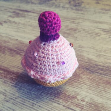Gehäkelter Cupcake mit rosa Creme, lila Häkelkirsche und Perlen auf Holztisch