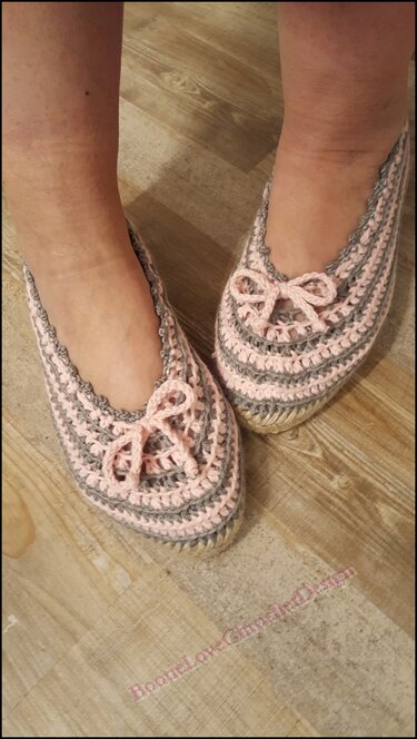 Füße mit gehäkelten Ballerina-Hausschuhen in Rosa und Grau mit Schleifen auf hellem Holzboden.