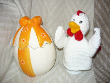 Weißer gehäkelter Huhn-Eierwärmer mit gelbem Schnabel und rotem Kamm neben einer orange verzierten Keramik-Eierschale