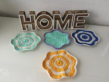 Vier gehäkelte, blumenförmige Untersetzer in Mint, Türkis, Blau und Gelb vor einem Holzschriftzug HOME auf weißer Oberfläche