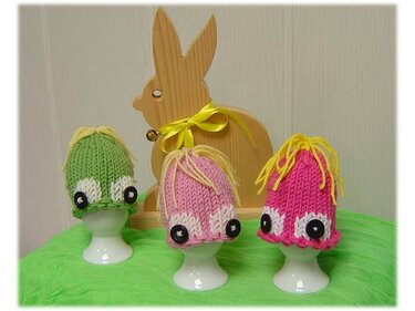Drei gestrickte Eierwärmer in Grün, Rosa und Pink mit Knopfaugen und gelben Wollhaaren auf weißen Eierbechern, dahinter ein Holzhase.
