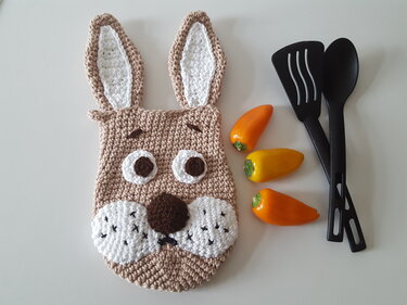 Gehäkelter Topflappen in Hasenform beige mit weißen Ohren und Schnauze, neben Kochutensilien und Paprika