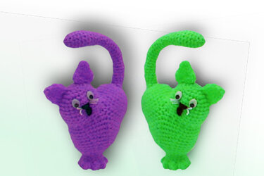 Zwei gehäkelte Amigurumi-Anhänger, einer lila, einer grün, mit Schwanz und Knopfaugen