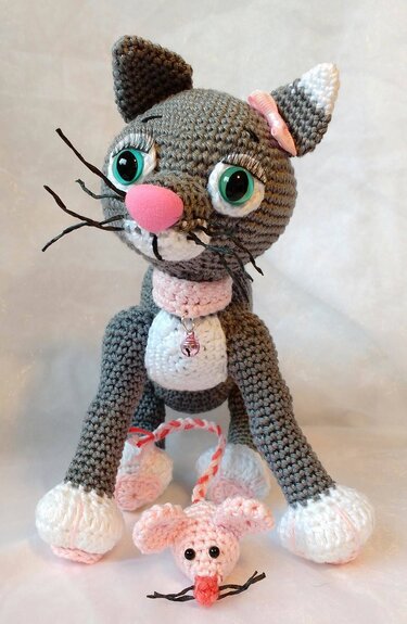 Gehäkelte graue Amigurumi-Katze mit rosa Nase, grünen Augen, weißen Pfoten und rosa Halsband, davor eine kleine rosa Häkelmaus an Kordel.