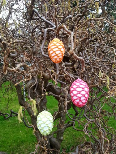 Drei mit Häkelnetz umhüllte Ostereier in Orange, Pink und Hellgrün, hängend an einem Baum mit gewundenen Ästen.