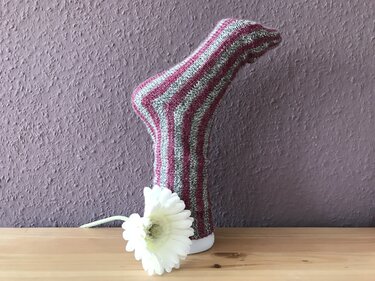 Gestreifte Stricksocke in Pink und Grau auf weißem Fußmodell mit weißer Blüte davor.