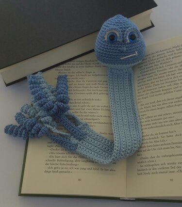 Gehäkeltes blaues Oktopus-Lesezeichen mit flachem Band und spiraligen Tentakeln auf offenem Buch