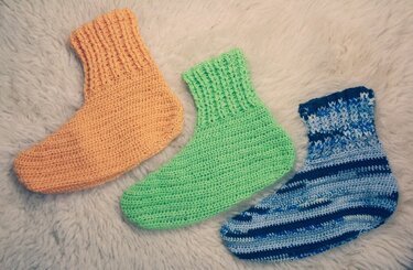 Drei gehäkelte Damensocken in Apricot-Orange, Hellgrün und Blau-Weiß auf weißem, flauschigem Hintergrund