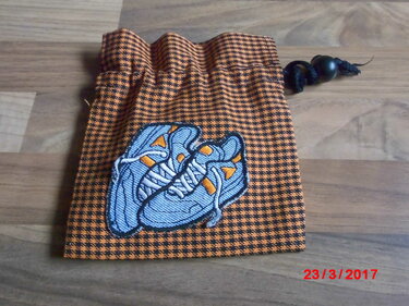 Orange-schwarzer Kordelzugbeutel im Hahnentrittmuster mit blauer Turnschuh-Stickerei auf Holzuntergrund.