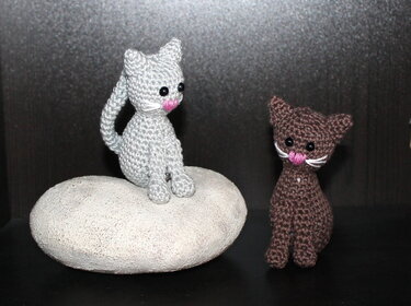 Zwei gehäkelte Amigurumi-Katzen (grau und braun), eine sitzt auf einem runden Stein, beide mit schwarzen Augen, rosa Nase und gestickten Schnurrhaaren