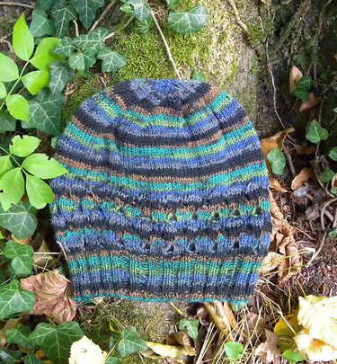 Striped knitted beanie in blue, green, brown and grey with textured stitch band, placed on moss and leaves.