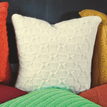 Weißes gestricktes Kissen mit geometrischem Reliefmuster, umgeben von bunten Kissen