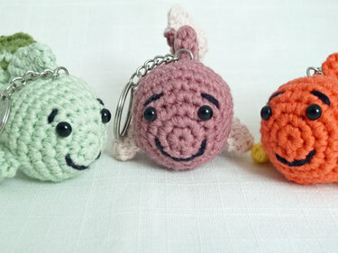 Nahaufnahme von drei handgehäkelten Amigurumi-Fisch-Schlüsselanhängern in Hellgrün, Rosa und Orange mit Metallringen.