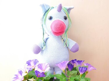 Hellblaues gehäkeltes Amigurumi-Einhorn mit pinker Schnauze und bunter Mähne vor lila Blüten