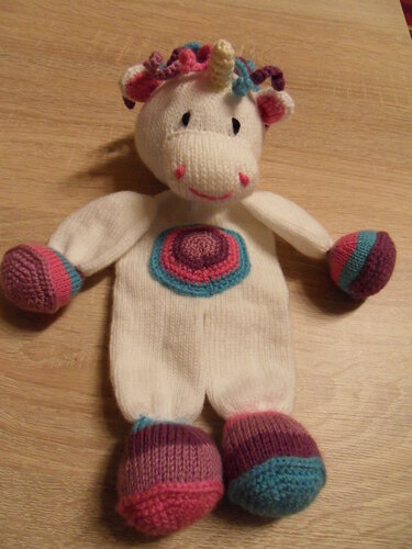 Gestricktes weißes Einhorn-Kuscheltier mit bunter Mähne, farbigen Händen und Füßen und rundem mehrfarbigem Muster auf dem Bauch, auf Holztisch liegend
