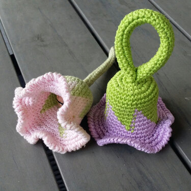 Zwei gehäkelte Babyrasseln in Glockenform, rosa-grün und lila-grün mit Greifschlaufe auf grauem Holztisch.