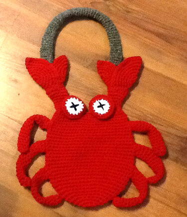 Rote gehäkelte Henkeltasche in Krabbenform mit grauem Henkel auf Holzuntergrund.