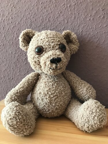 Beigefarbener gehäkelter Teddybär mit großen Augen, sitzt auf einem Holztisch.
