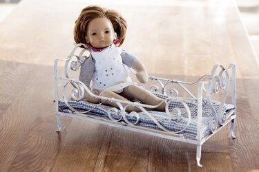 Puppe mit weißem Kleid und rotem Halstuch sitzt auf weißem, verziertem Miniaturbett mit kariertem Polster auf Holzfußboden