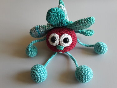 Gehäkeltes Amigurumi mit rotem Körper, weißen Augen, türkisfarbenen Blättern und vier Kugelbeinen
