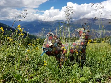 Genähtes Kuscheltier in Katzenform aus geblümtem Stoff mit Knopfauge und blauer Garnschnur in grüner Bergwiese vor Bergen