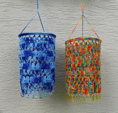 Zwei gehäkelte Hängehüllen, blau und mehrfarbig, mit Perlenfransen vor heller Wand